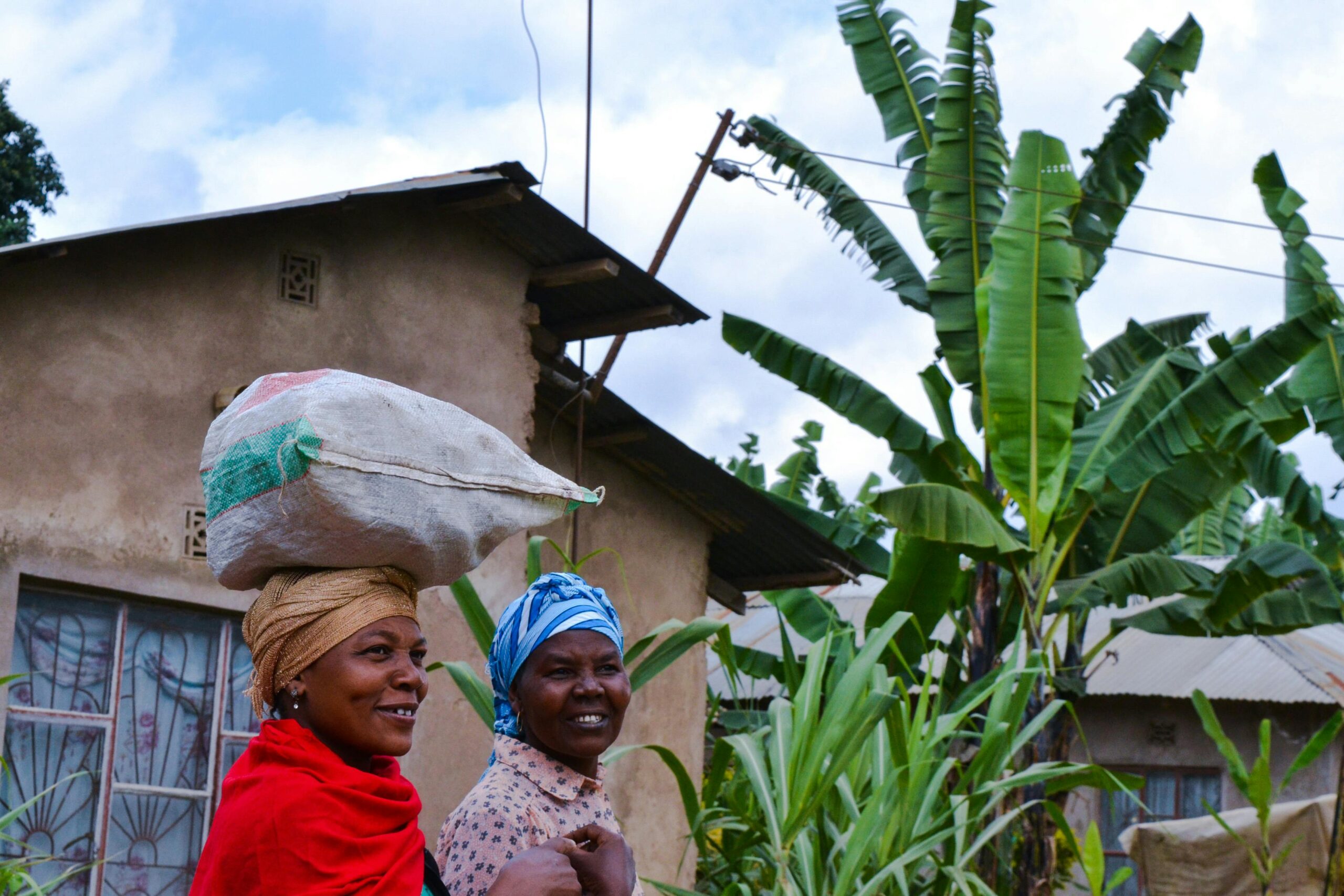 Projetos Smiling women in Arusha, Tanzania carrying a sack over traditional attire in a village setting.